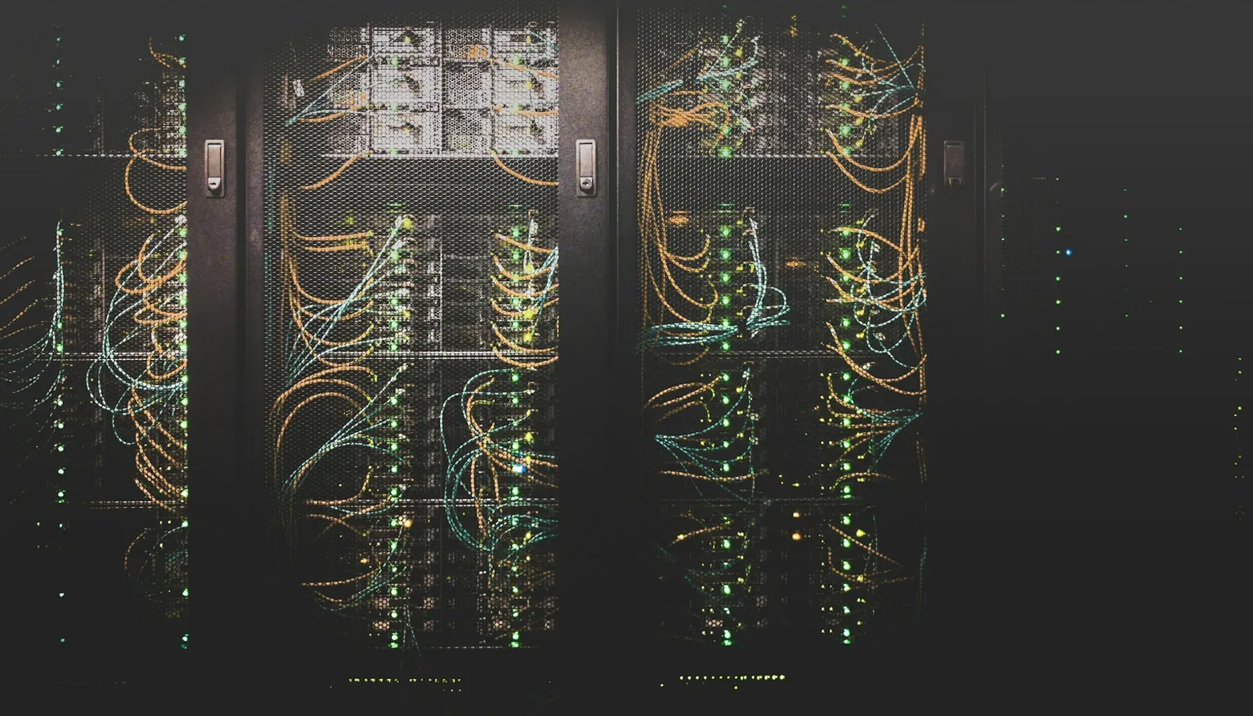 Photograph of a dark data centre aisle: server racks behind mesh doors, threaded with orange and teal cables and lit by rows of small green status lights